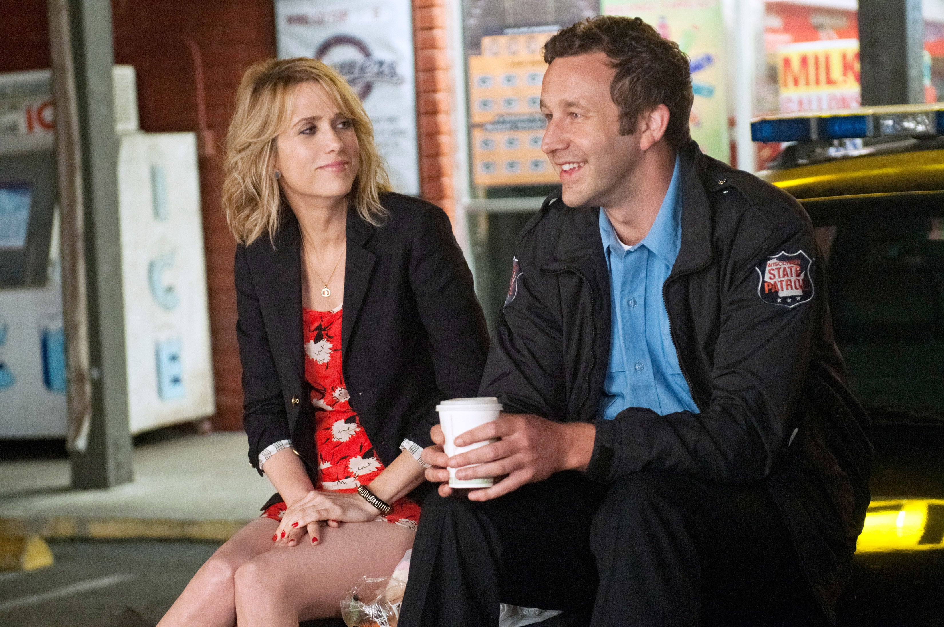 Two people sitting on a car hood, talking and smiling. One wears a floral dress with a black blazer; the other is in a police uniform with a coffee cup