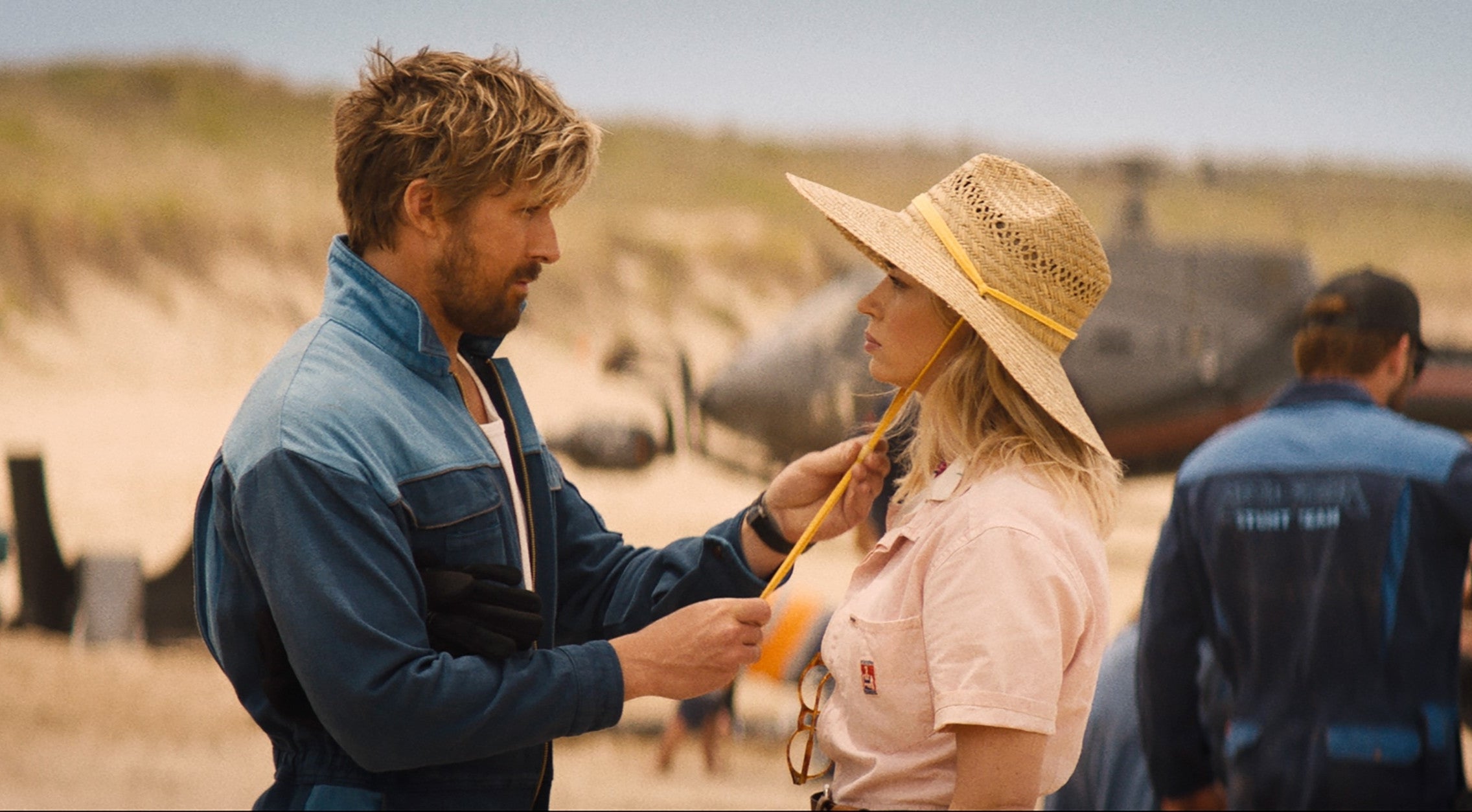 A man and woman face each other on a beach; he adjusts her jacket. She wears a straw hat. Others work in the background