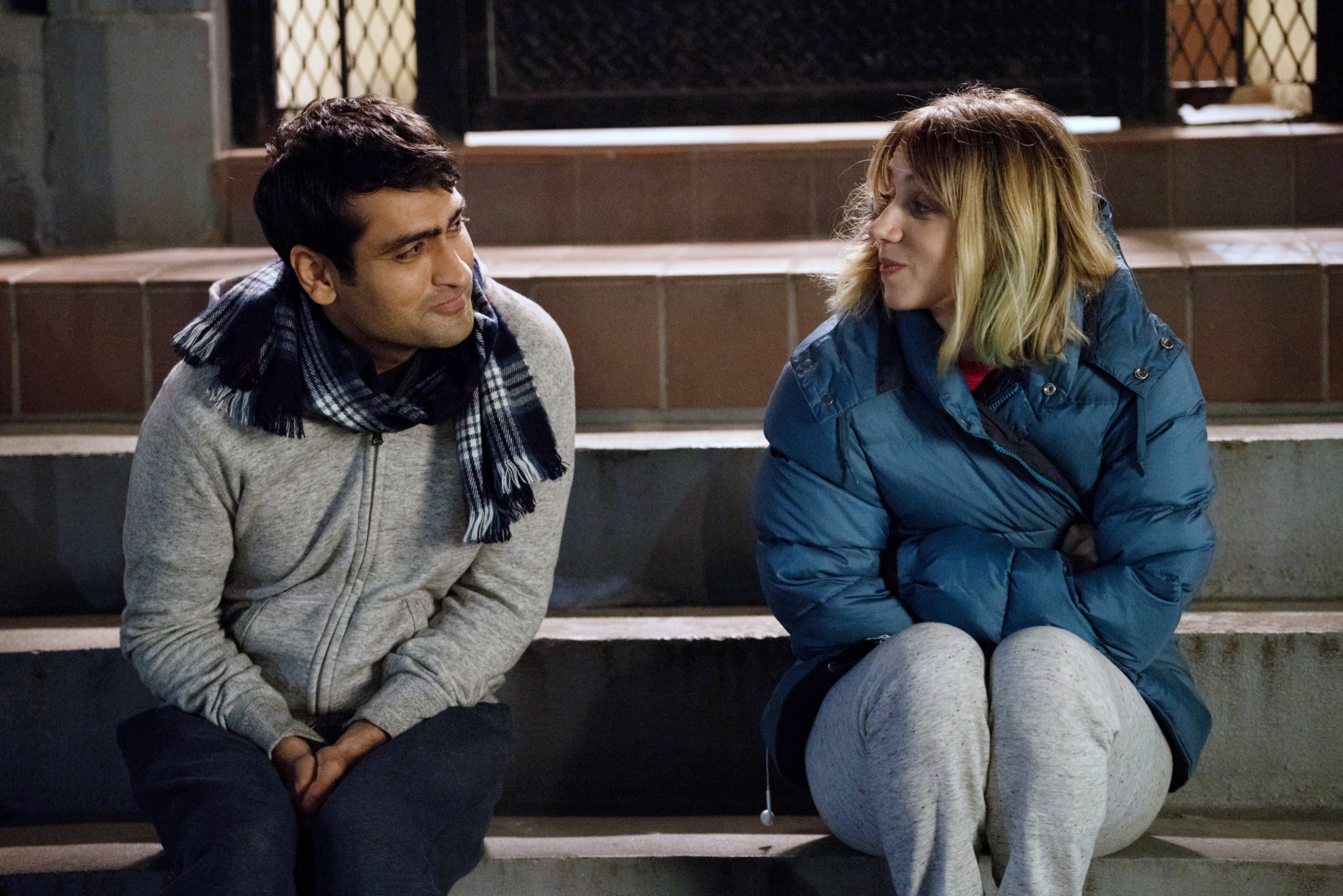 Two people sit on outdoor steps, talking warmly. The person on the left wears a scarf and jacket, while the person on the right is in a puffy coat