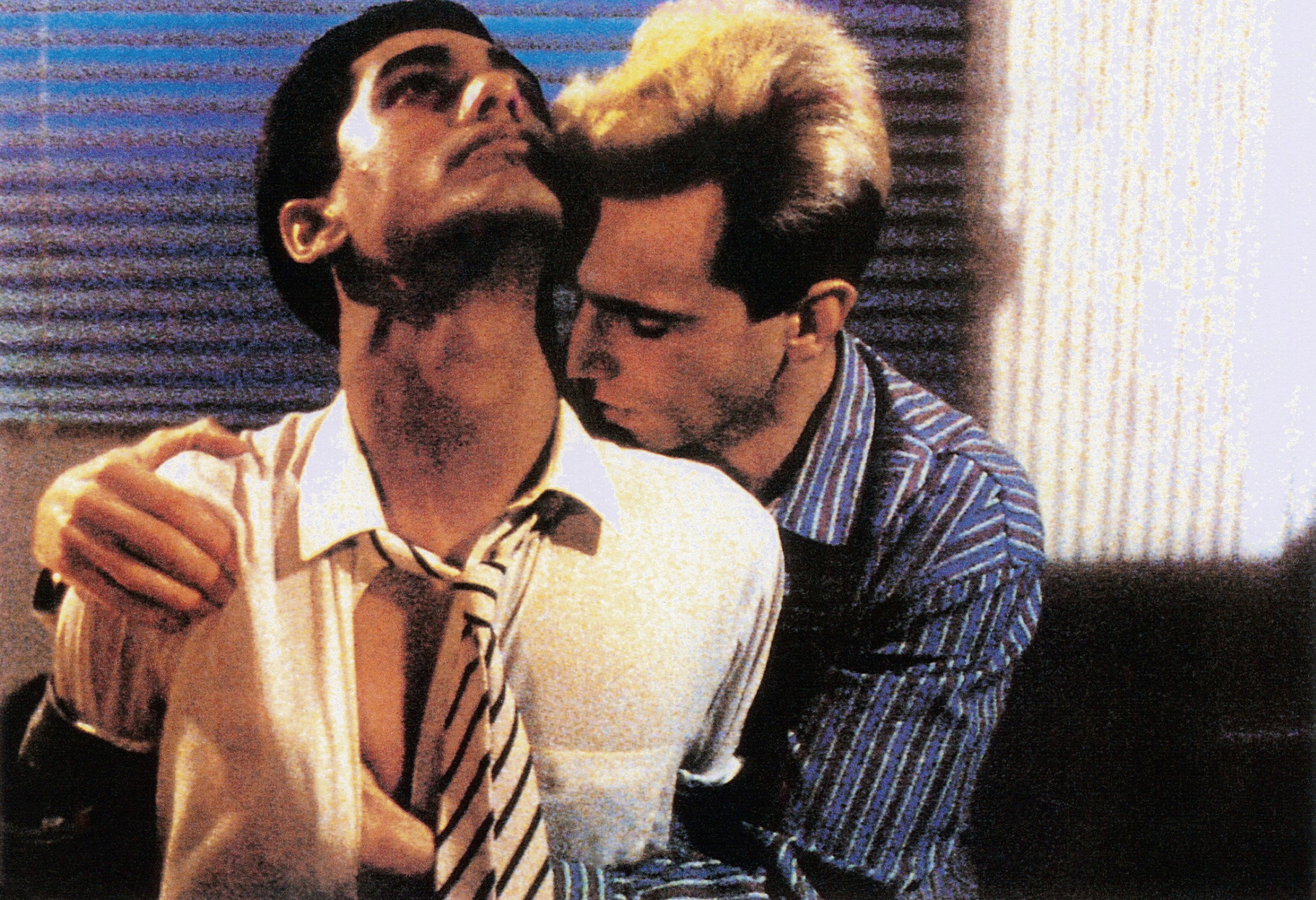 Two individuals in an intimate pose, one wearing a shirt and tie, the other in a striped shirt, in a dimly lit setting