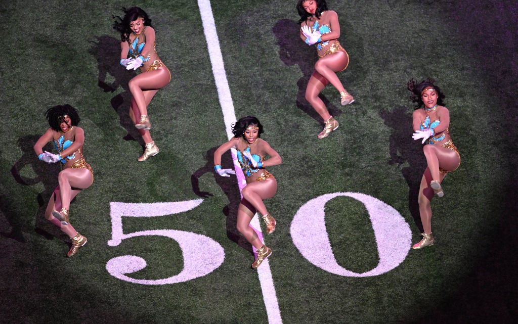 Five performers in synchronized gold and teal outfits dance energetically on a football field over the number 50