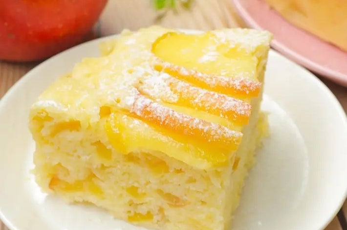 皿に乗ったアップルケーキのスライス。表面にリンゴが並んでいる。背景にリンゴと別のケーキが見える。