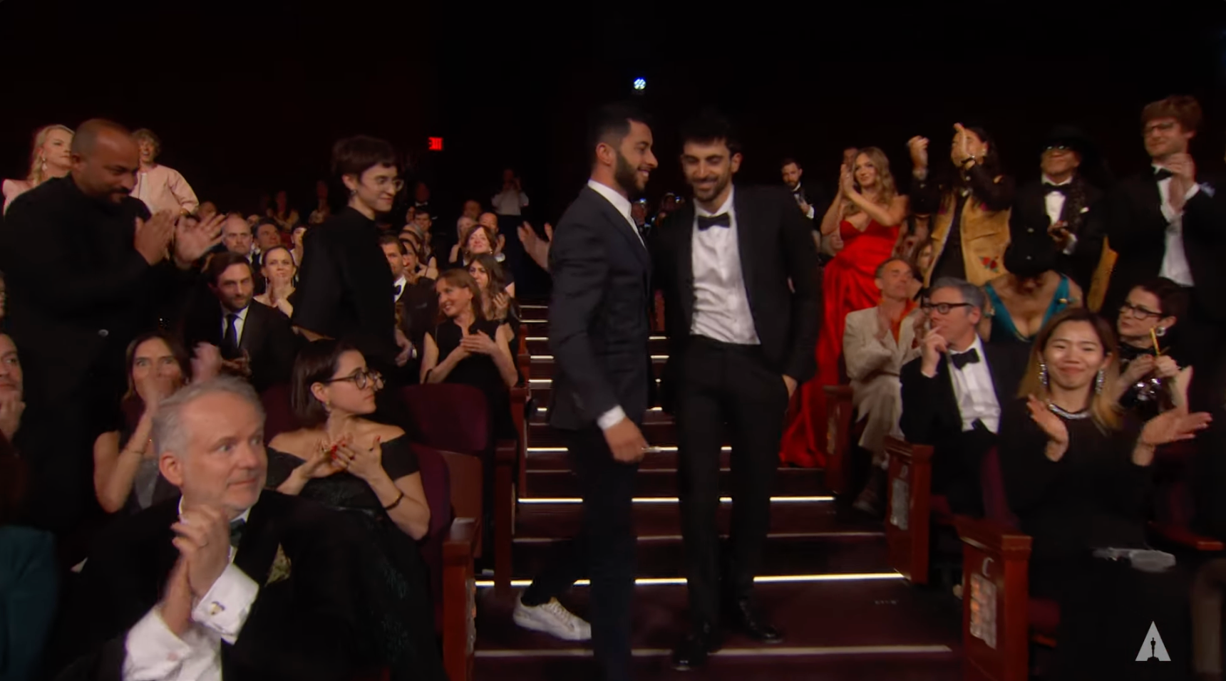 Audience applauding two people standing in a theater aisle; one wearing a suit with sneakers, the other in a tuxedo
