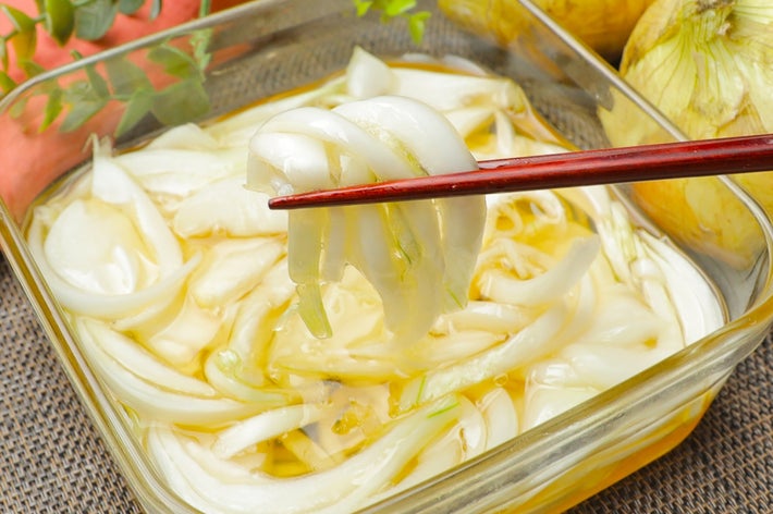 玉ねぎのピクルスがガラス容器に入れられており、箸で持ち上げられている。後ろには生の玉ねぎが見える。