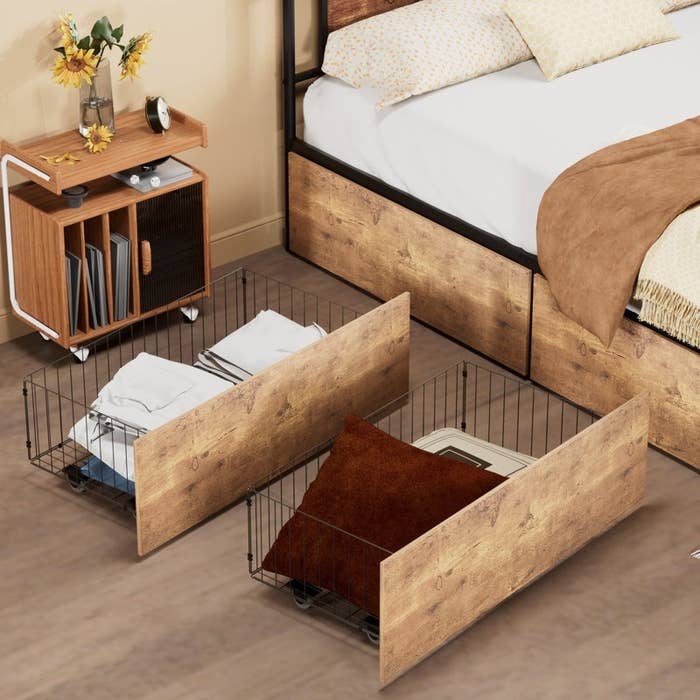 Under-bed storage bins with wooden fronts, metal sides, and wheels, showing folded clothes and a plush pillow inside. Bedside table holds decorative items