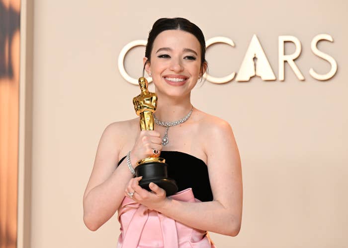 Person in elegant evening gown holding an Oscar statue, posing in front of a backdrop with the word &quot;OSCARS.&quot;