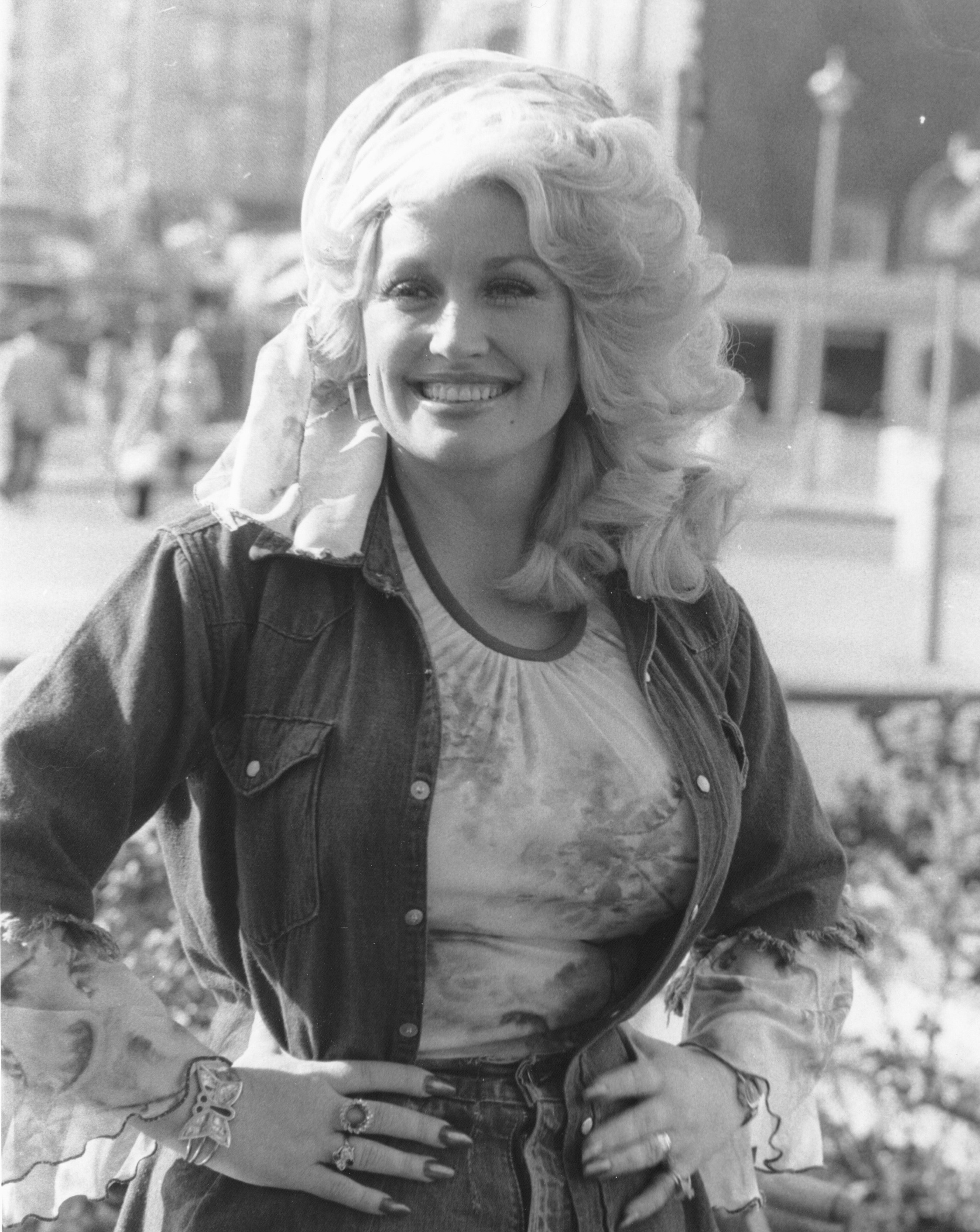 Person in a denim jacket and patterned blouse smiling outdoors with hands on hips