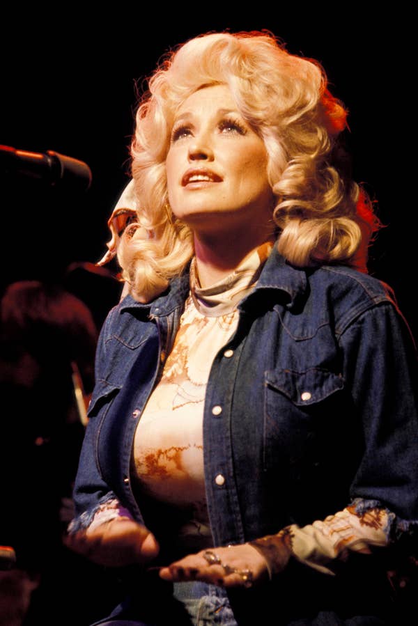 Person with voluminous hairstyle in a denim shirt and patterned top, seated on stage, looking upwards during a performance