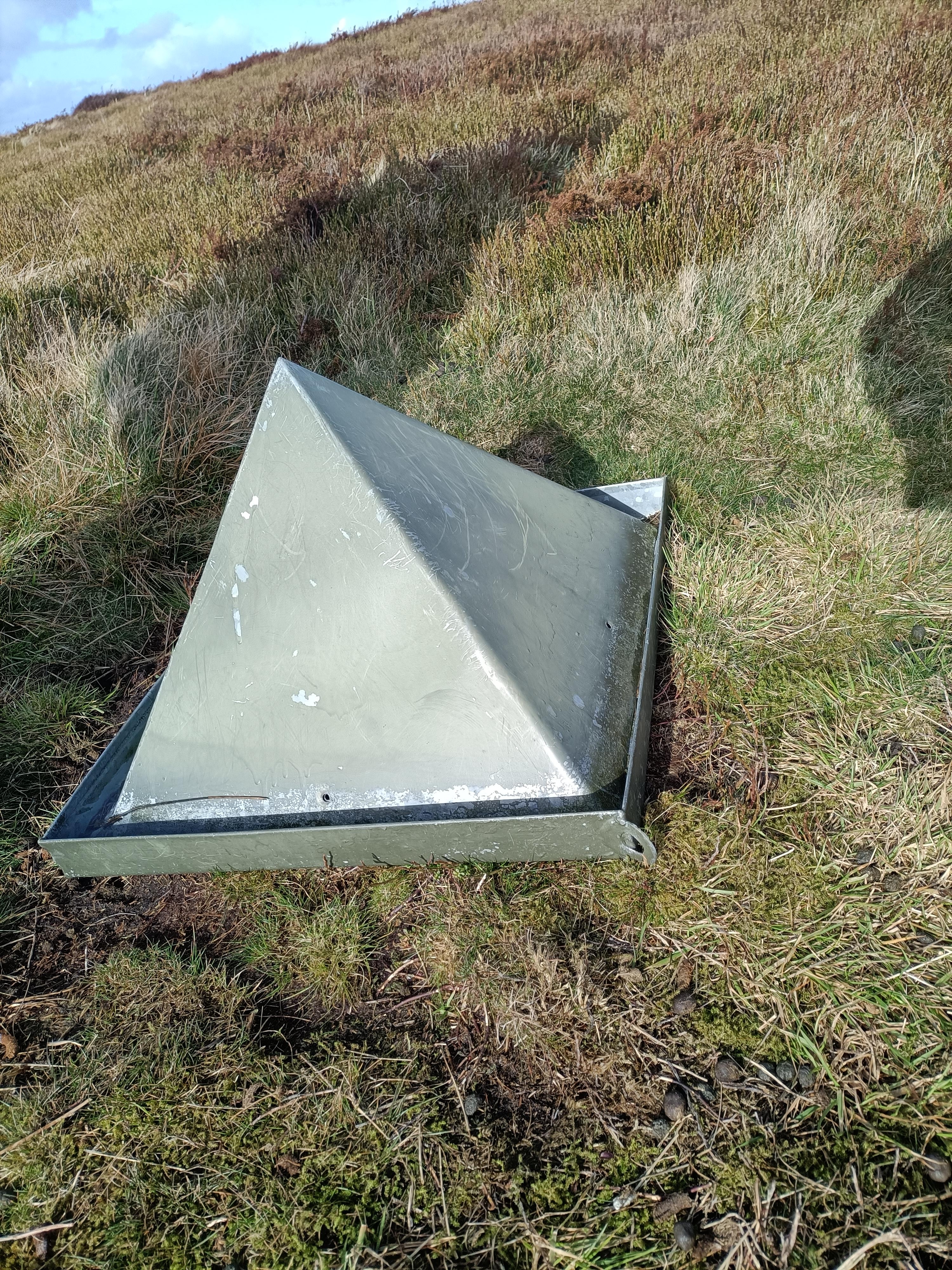 Metal pyramid-shaped object lying on grassy hillside, surrounded by shadow and natural elements