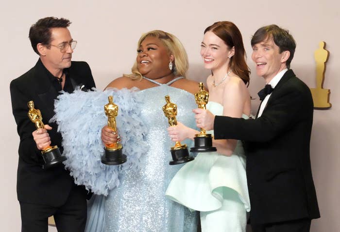 Four people on a red carpet smiling and holding awards; two wear elegant gowns and two wear suits