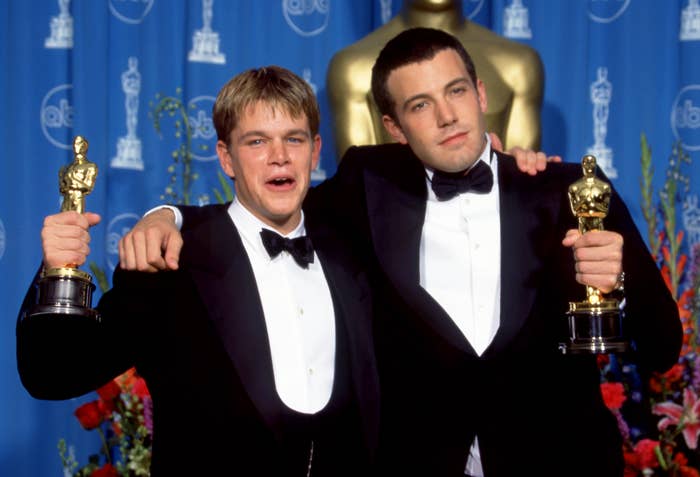 Two men in tuxedos holding Oscars, posing with smiles and arms around each other, in front of an Oscars backdrop