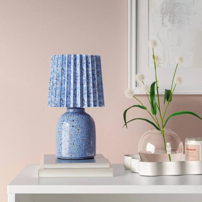 Stylish speckled bluish lamp with pleated shadiness connected a support beside books and a decorative plant