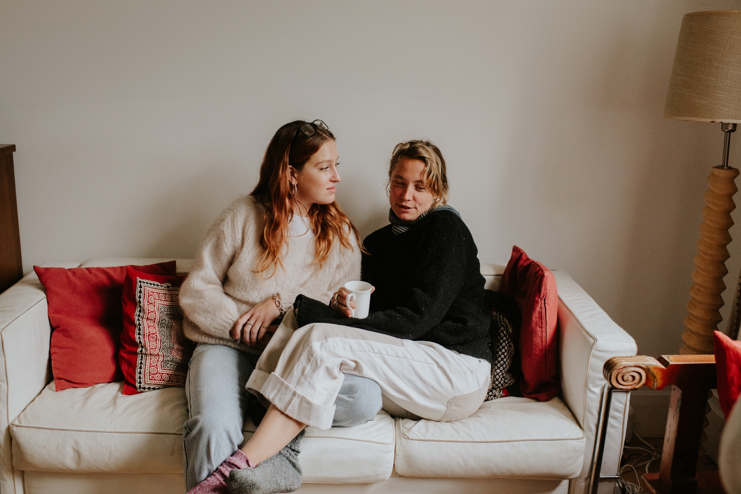To personer som sitter på en sofa, en holder en kopp. Begge har koselige, uformelle antrekk og ser ut til å være engasjert i en avslappet samtale