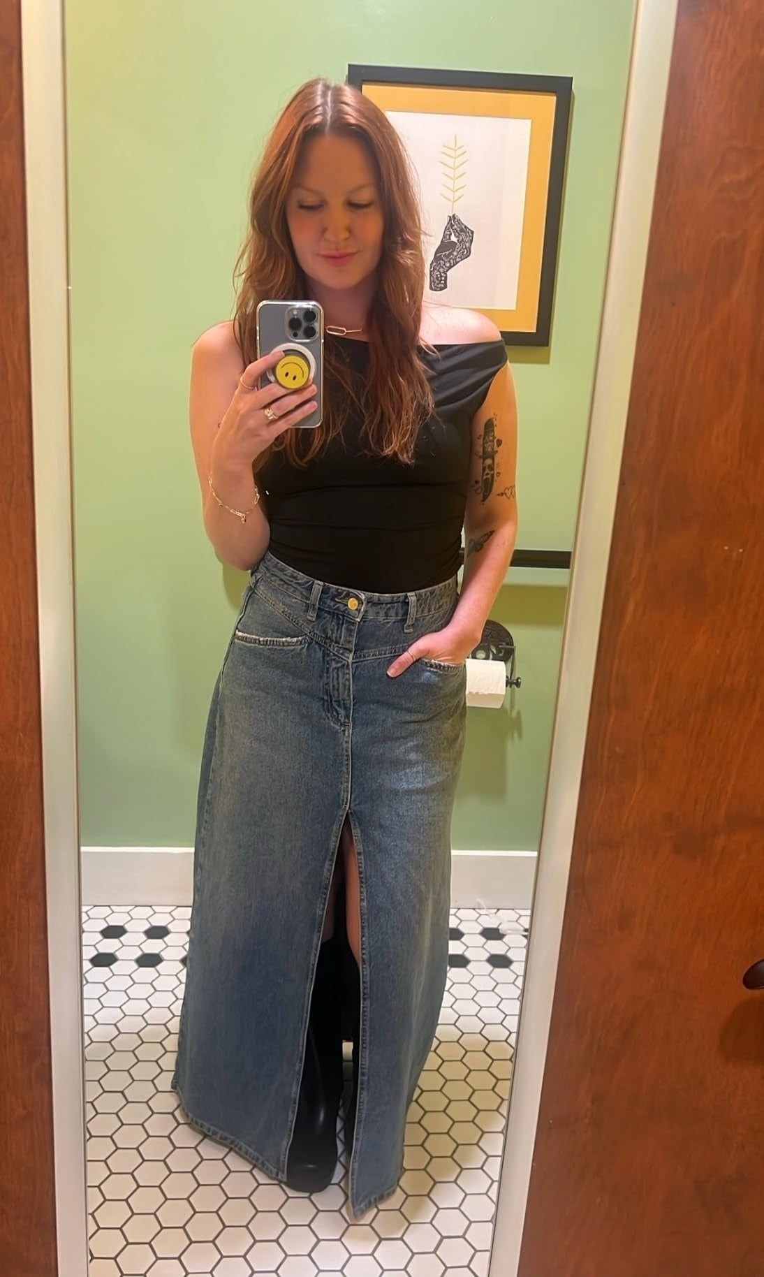 Orang yang mengambil selfie cermin di kamar mandi, mengenakan atasan di atas bahu dan rok denim panjang dengan celah depan