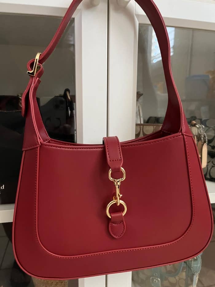 Tas tangan merah dengan gesper emas ditampilkan di rak, cocok untuk penggunaan sehari -hari yang penuh gaya