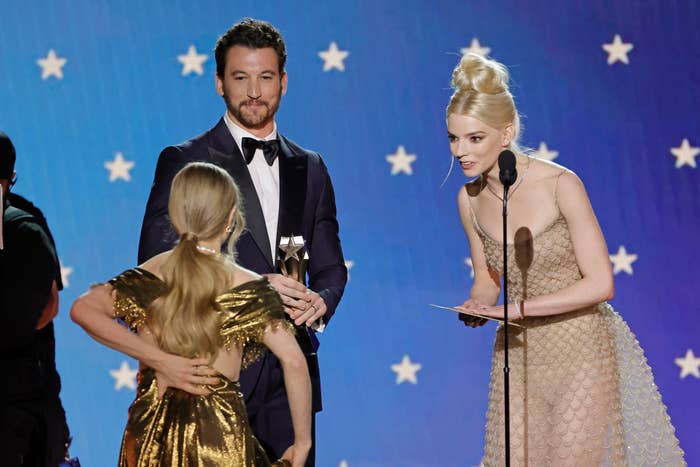 Amanda Seyfried, Miles Teller, and Anya-Taylor Joy on stage at an award show. One man in a tuxedo holds an award; two women in elegant dresses face him, one speaking into a microphone