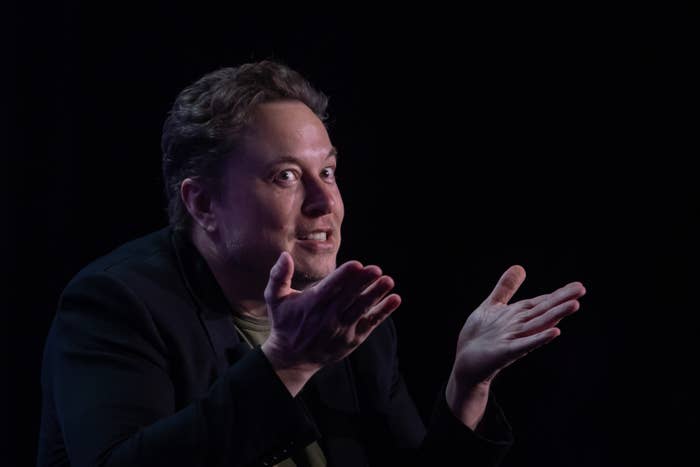 Elon Musk gesturing with hands open, wearing a casual blazer and shirt, against a dark background