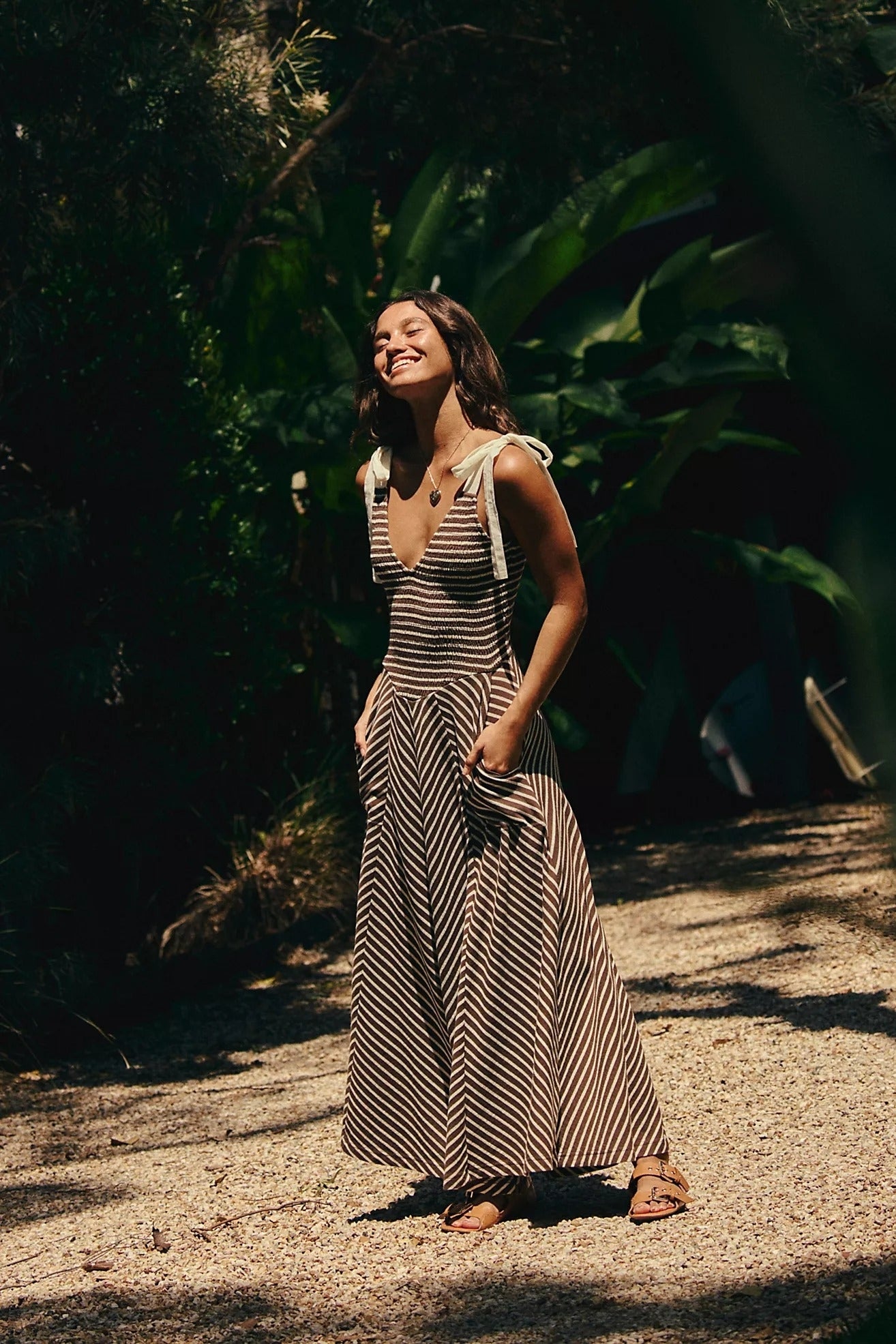 Person outdoors wearing a striped sundress with tie straps, standing on a gravel path, surrounded by lush greenery