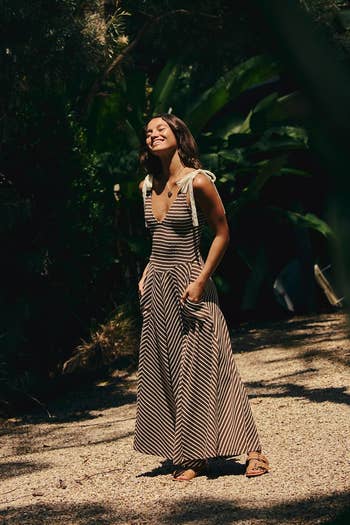 Person outdoors wearing a striped sundress with tie straps, standing on a gravel path, surrounded by lush greenery