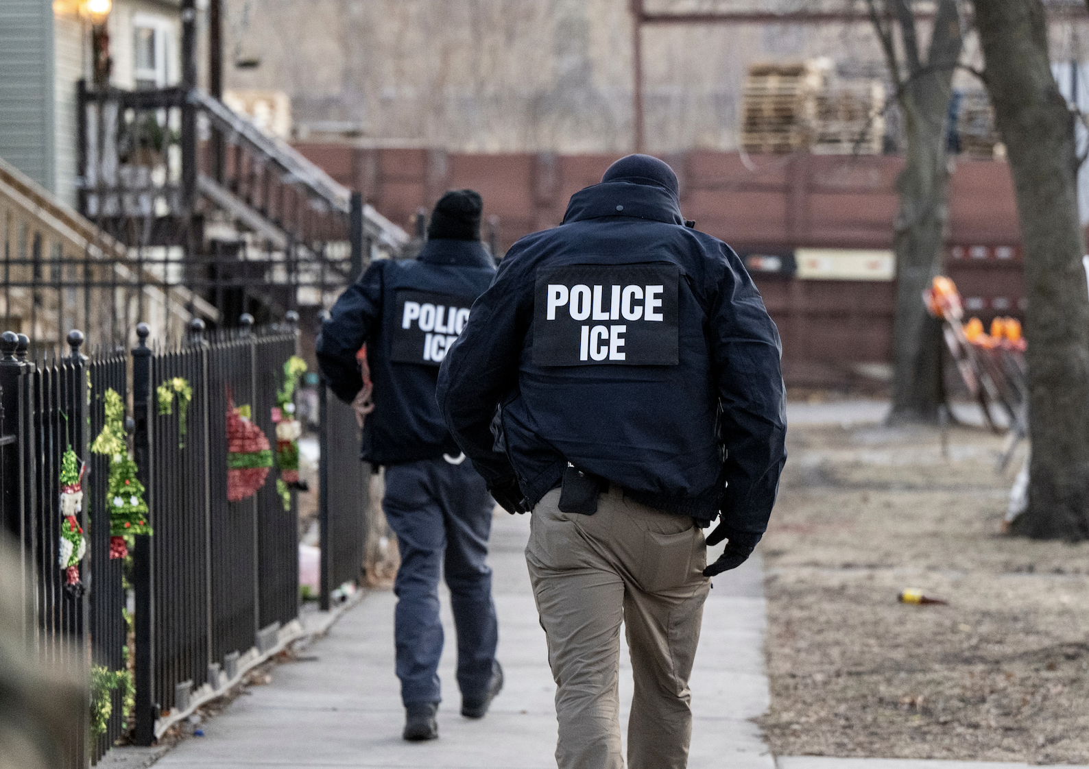 Two ICE officers in jackets with "POLICE ICE" on the back walk down a residential sidewalk near a fenced area
