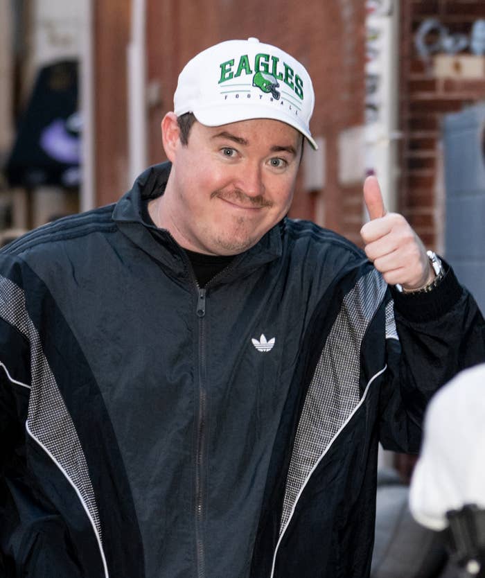 Shane Gillis in a cap and tracksuit smiling and giving a thumbs-up gesture