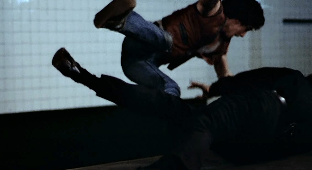 A person in jeans jumps towards another on the ground in a dramatic, action-packed scene, suggesting a fight