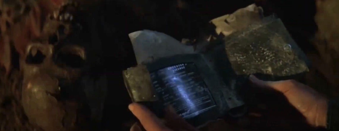 Person holding a futuristic, weathered device in a dark setting, displaying digital information. Skulls are faintly visible in the background