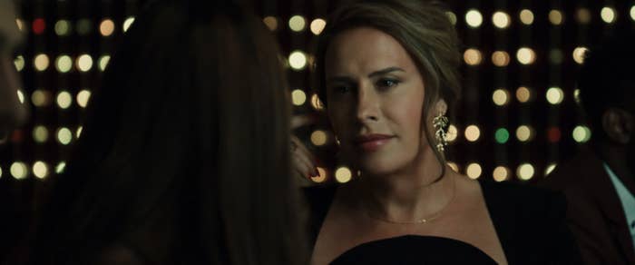 Karla Sofia Gascon in an elegant black outfit with statement earrings is engaged in conversation in a dimly lit setting with sparkling lights in the background