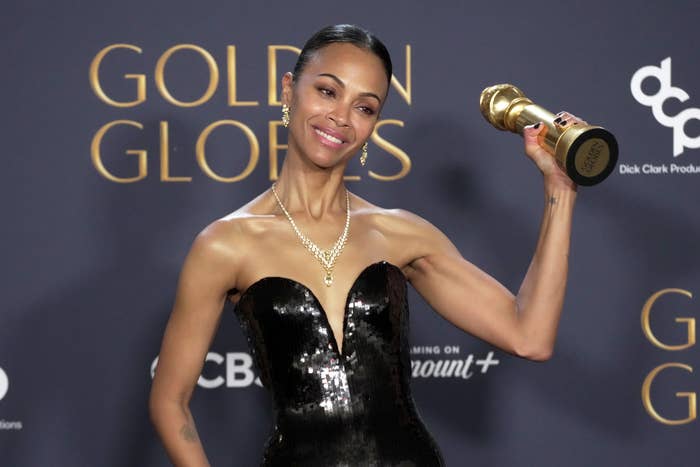 Zoe Saldana holding a Golden Globe award, wearing a strapless, sequined dress and elegant necklace, smiling at the camera