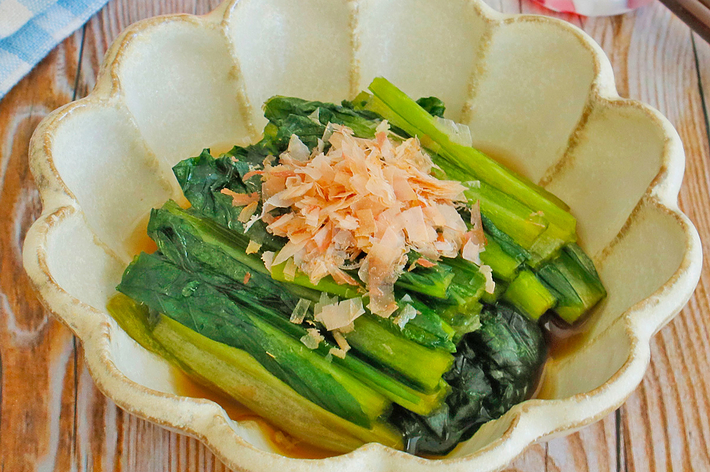 お浸しが椀に盛られており、上に鰹節が飾られています。背景には箸と花が置かれています。