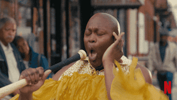 Person in an extravagant yellow outfit passionately sings into a microphone on a street, surrounded by buildings. Netflix logo visible