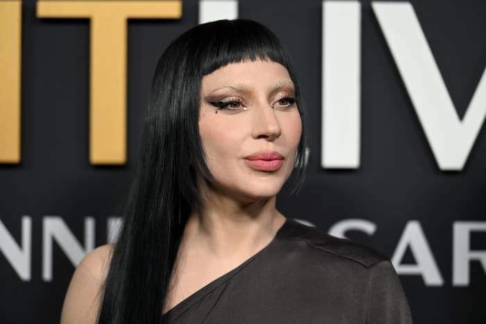 Gaga with long dark hair and asymmetrical bangs on a red carpet, wearing an elegant one-shoulder dress