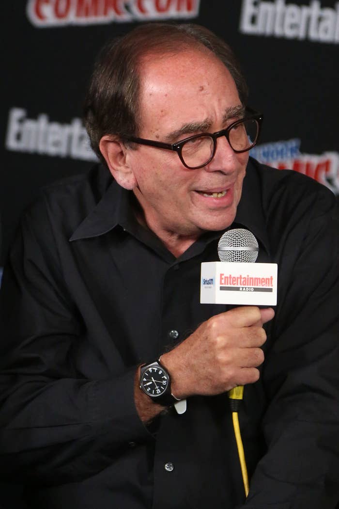 Man speaking into a microphone at a Comic Con event, wearing glasses and a dark button-up shirt