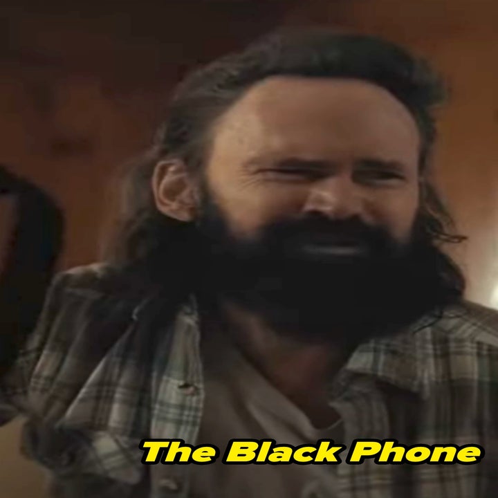 Jeremy Davies in The Black Phone with a beard and long hair, wearing a plaid shirt, holds up a belt, appearing frustrated in a rustic setting