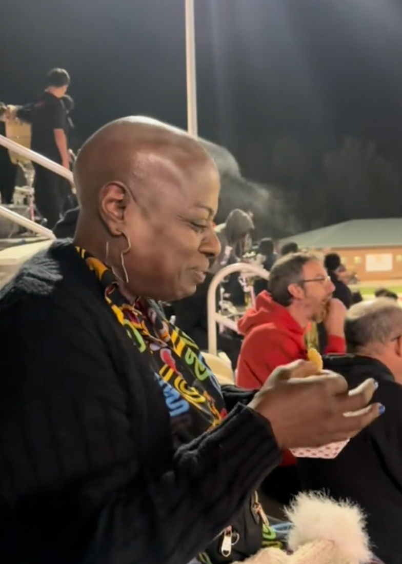 Personen smiler og ser på telefonen sin mens han sitter på et stadion med mennesker i bakgrunnen