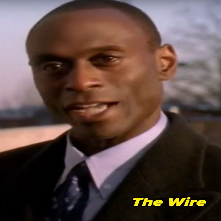 Lance as Daniels in the Wire in a formal coat and tie speaks outdoors, with blurred trees in the background