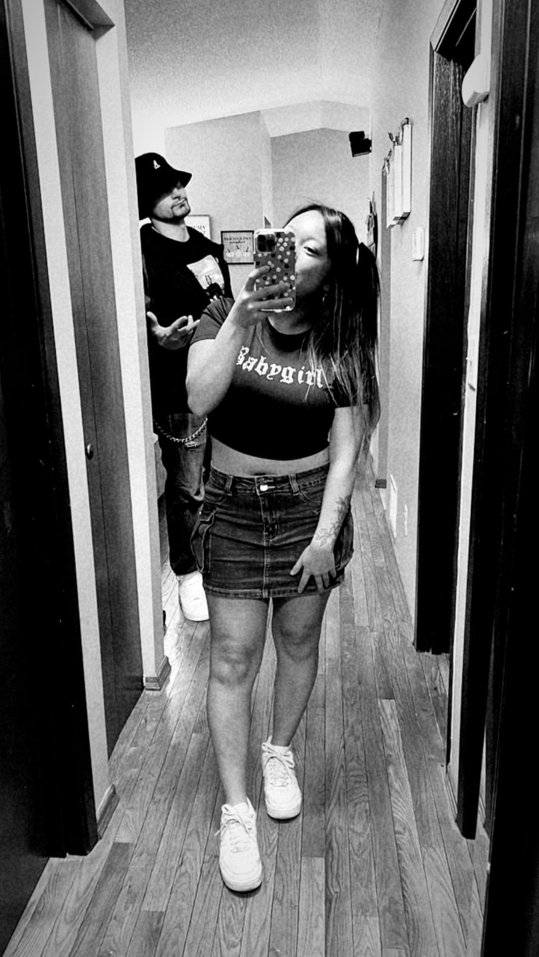 Dua orang di lorong, satu mengambil selfie cermin. Orang di depan mengenakan atasan dan rok denim. Orang di punggung mengenakan pakaian kasual