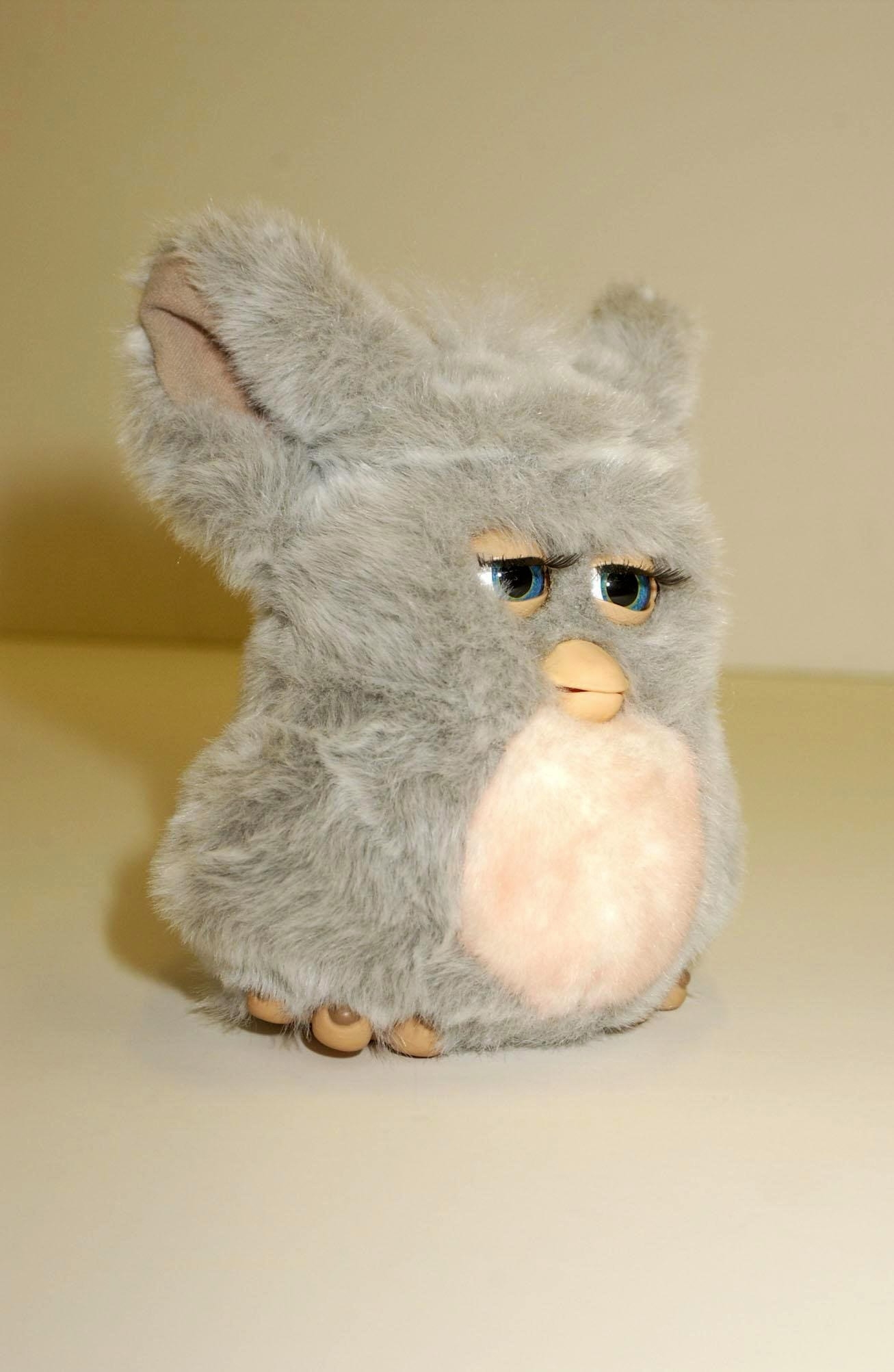Furby toy with grey fur and a white belly, featuring expressive eyes and large ears, posed against a plain background