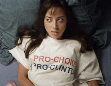 Aubrey's character lying on a bed, wearing a T-shirt with the text "Pro-Choice, Pro-Clinton"