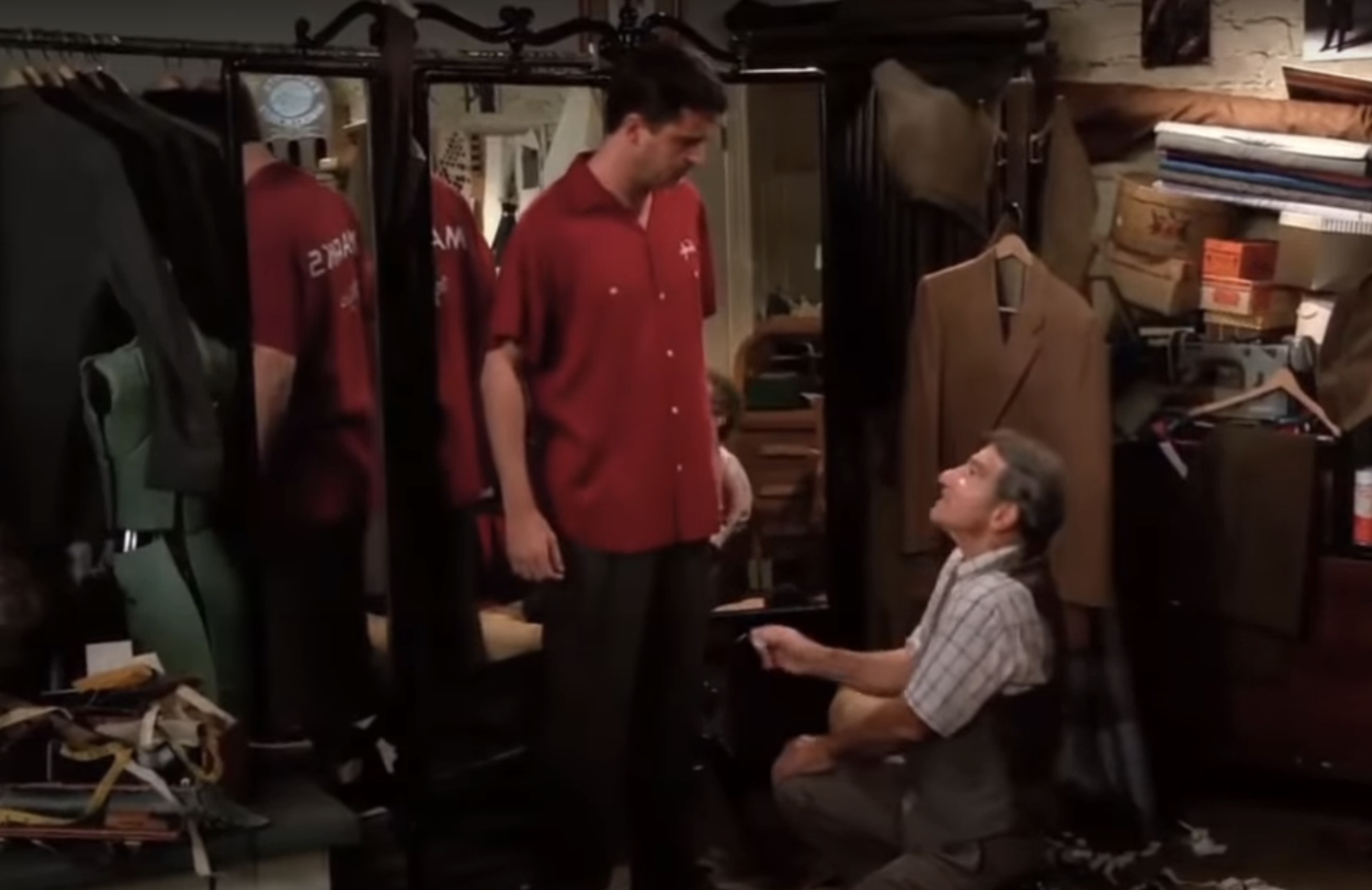 A man stands in front of a mirror wearing a red shirt, while a tailor kneels, adjusting the fit of his pants in a cluttered workshop