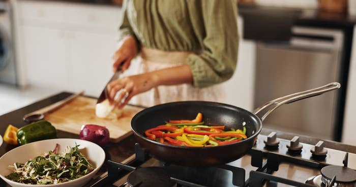 Person chopping an onion on a wooden board next to a stove with a pan of sautéing bell peppers and a salad bowl in the foreground