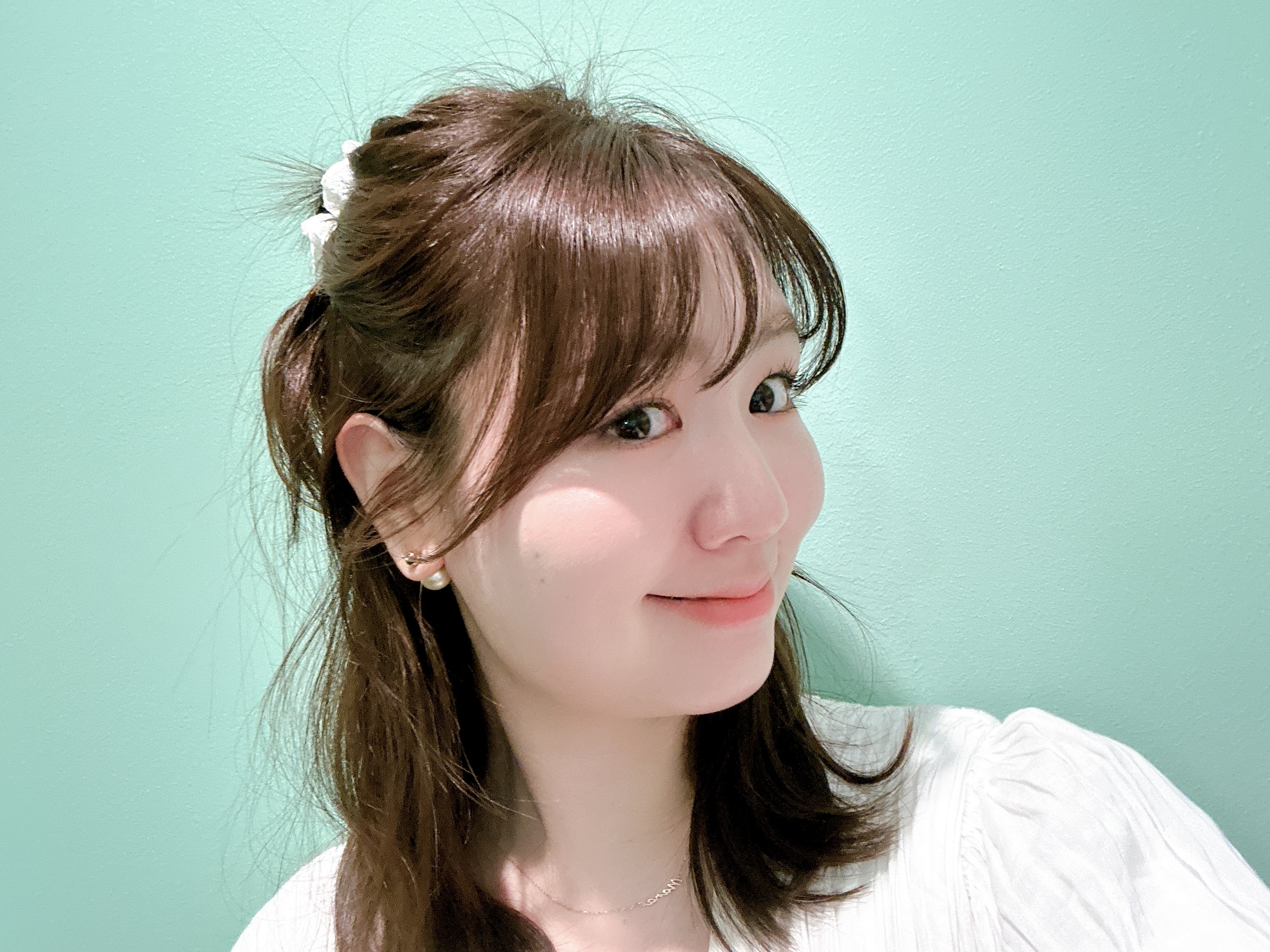 女性がカメラに向かって微笑んでいます。髪はハーフアップで、白いアクセサリーをつけています。