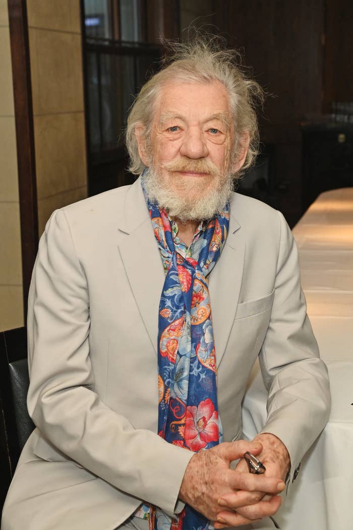 An elderly individual with a beard wearing a light suit and floral scarf sits at a table, hands clasped, looking at the camera
