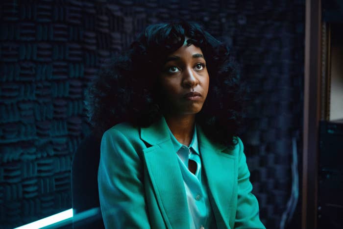 Person in a recording studio wearing a stylish green blazer and blouse, looking thoughtful. Soundproofing panels are visible in the background