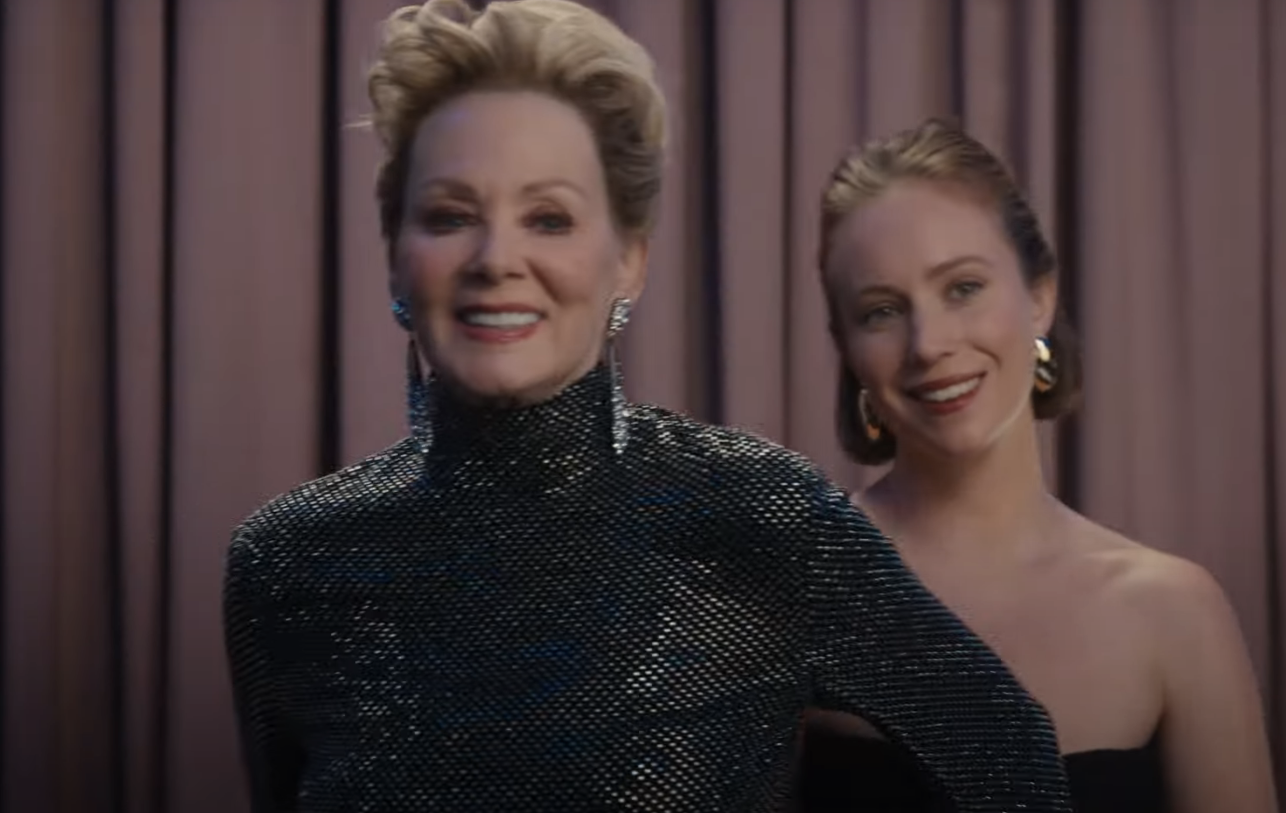 Deborah and Ava smiling in front of draped curtains; one in a sparkling turtleneck dress, the other in a strapless outfit with hoop earrings