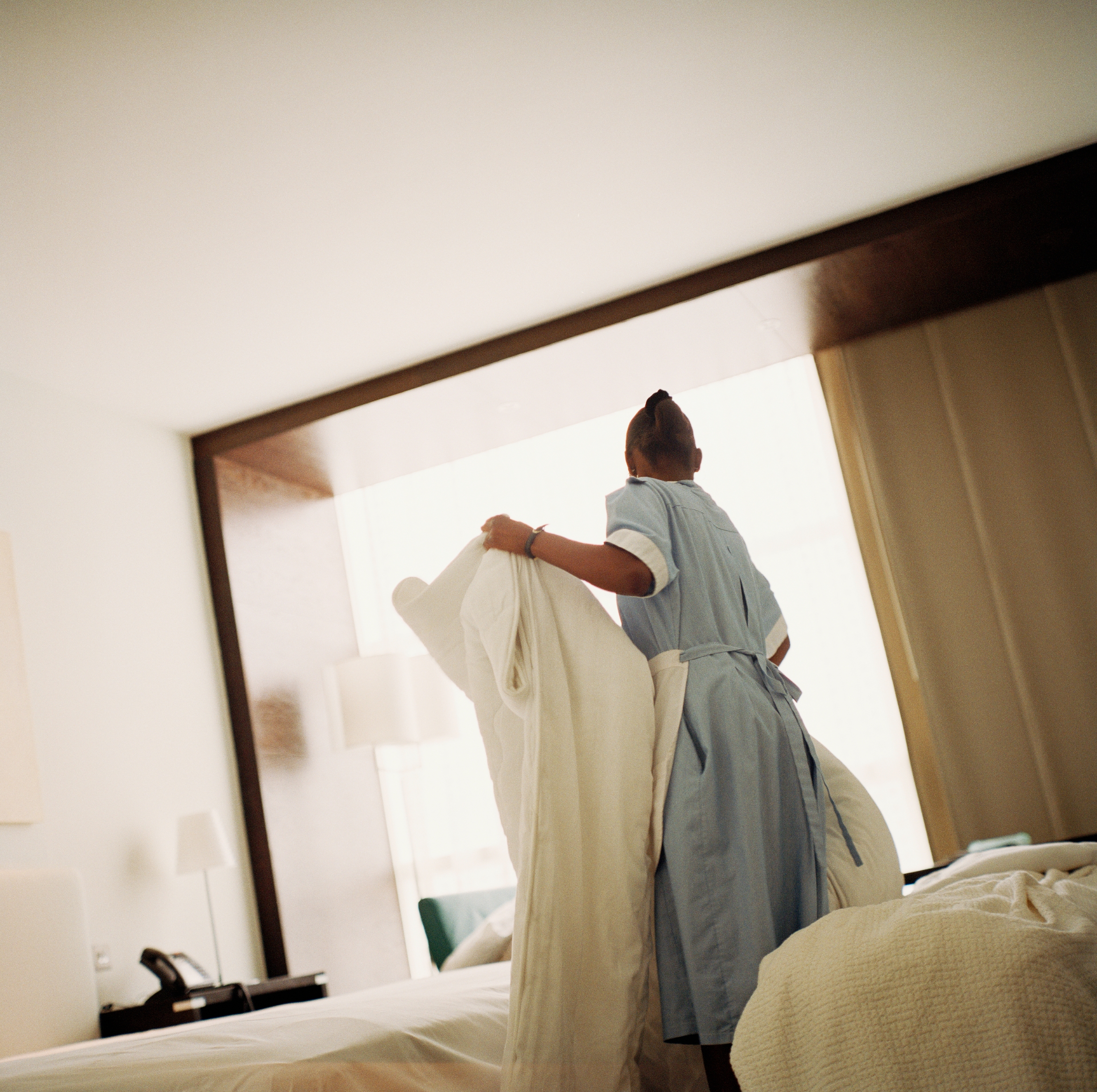 Hotellarbeider i uniform lager en seng i et solfylt rom, og arrangerer ark med omhu