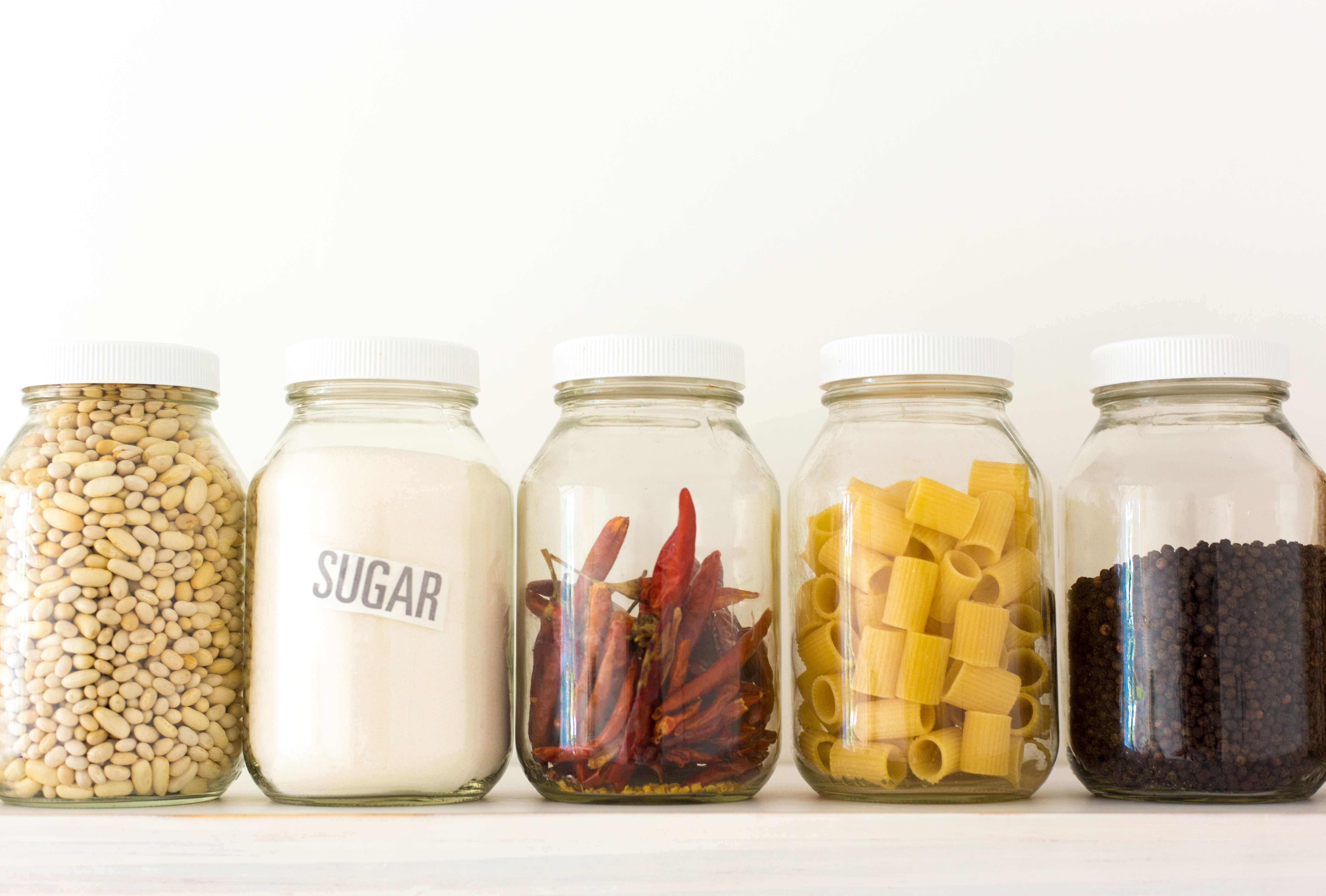 Five glass jars with white lids holding beans, sugar, red chili peppers, pasta, and black pepper are arranged in a row on a countertop
