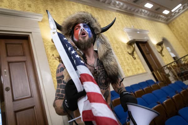 Man in fur hat with horns and flag face paint holding a U.S. flag and megaphone in the Senate chamber