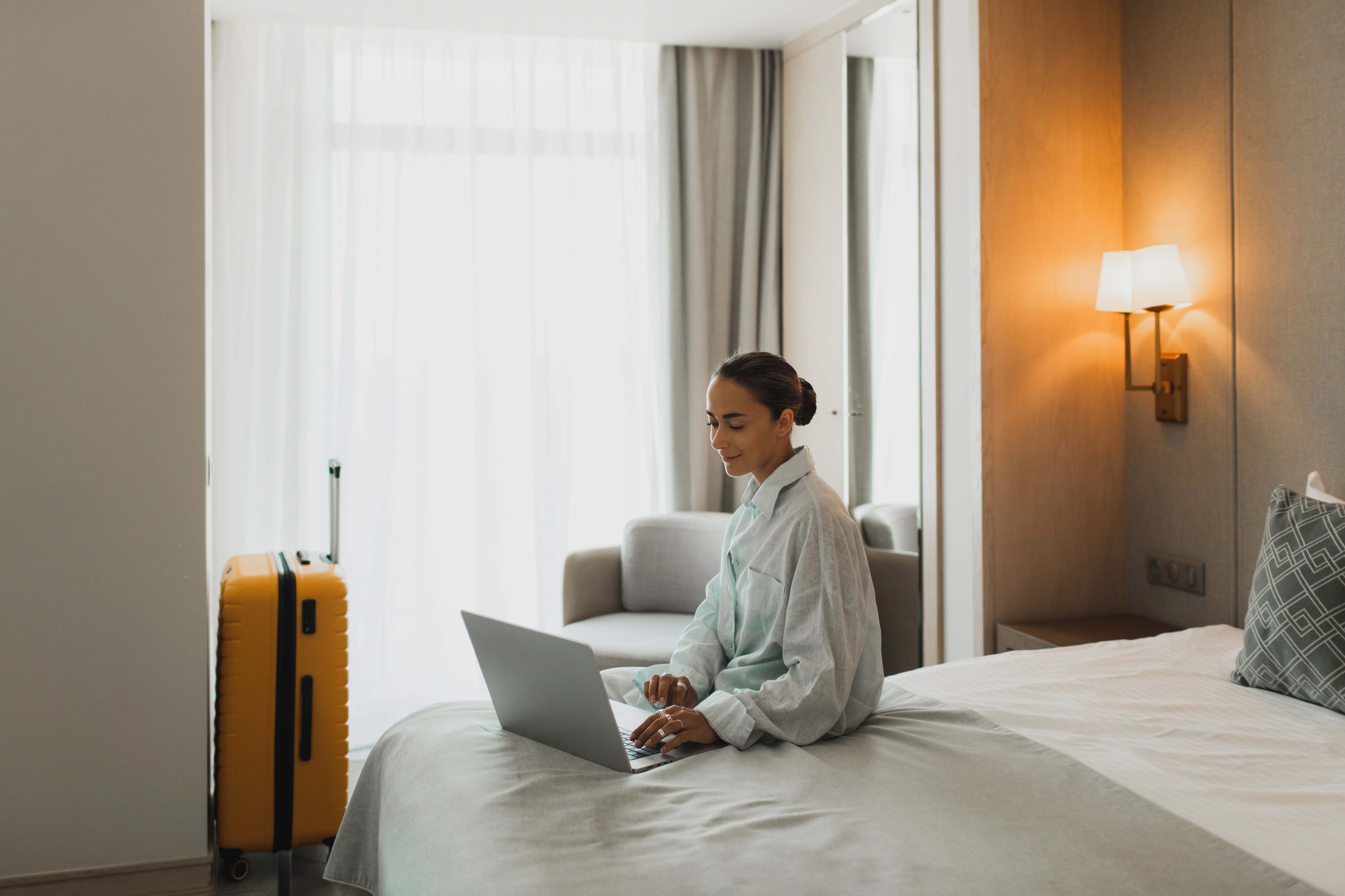 Person som sitter på en hotellseng ved hjelp av en bærbar PC, en gul koffert i nærheten, i et godt opplyst rom med moderne dekor