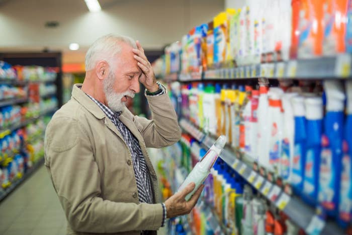 Elderly man in a store aisle, looking confused while holding a product, surrounded by various cleaning supplies on shelves